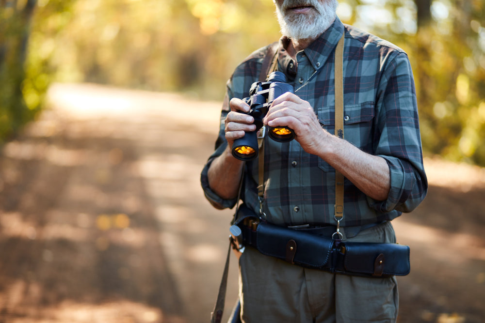 Older man in flannel shirt uses binoculars to scout for deer in the woods